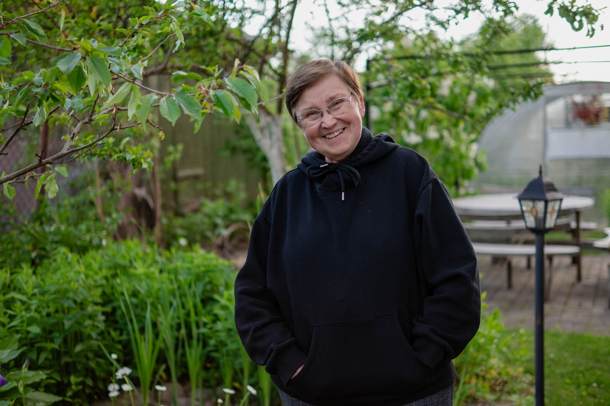 Елена стоит во дворе, улыбается, руки в карманах; зелень, стол, теплица.