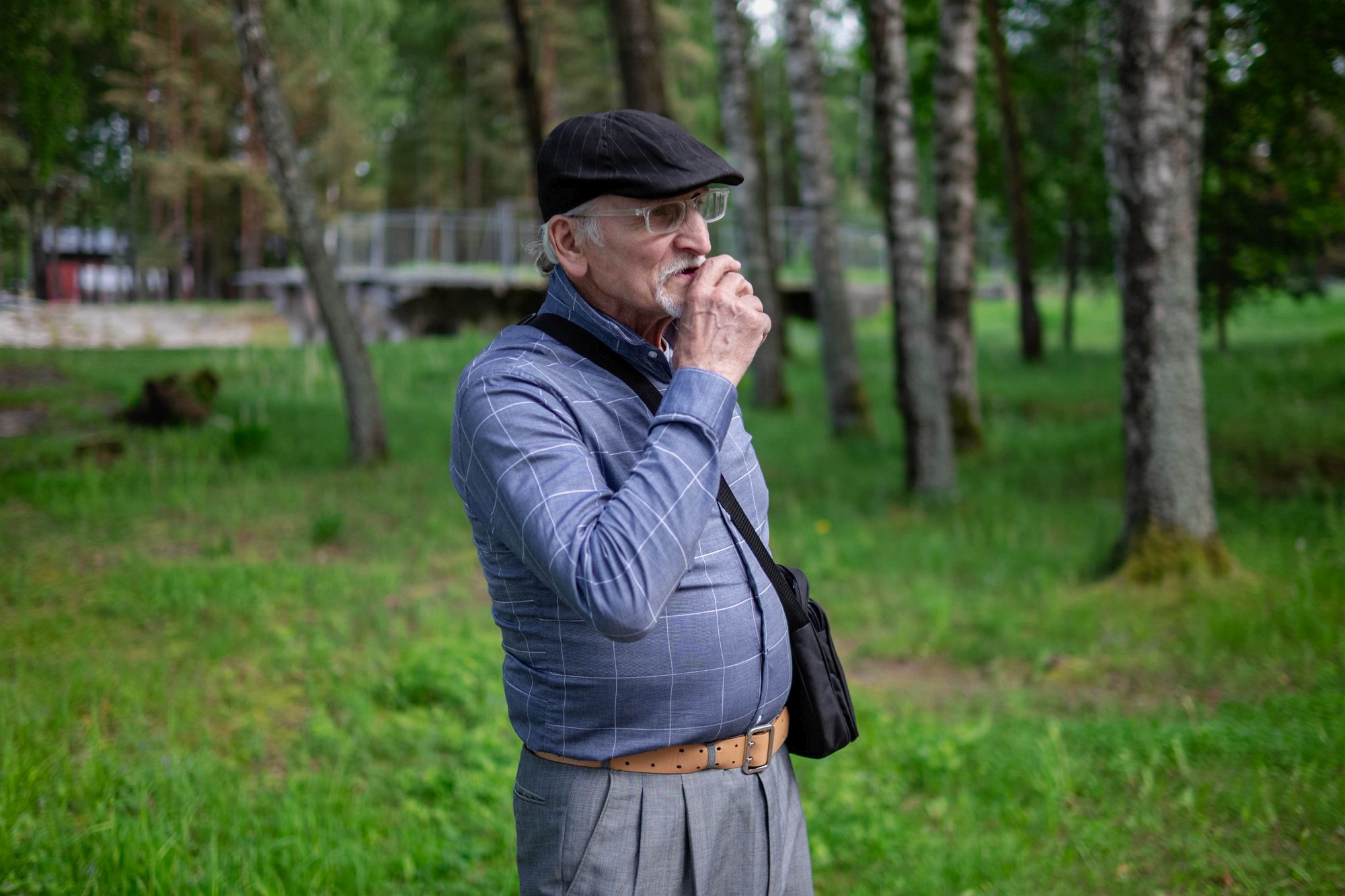 Юрий среди деревьев в зелёной зоне, держит руку у лица, будто делает паузу в разговоре. На заднем плане мостик и вода.